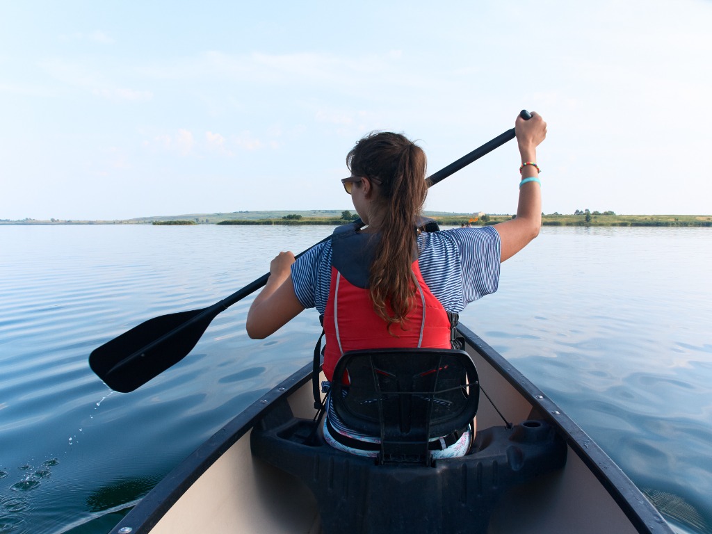 Señora joven con el pelo moreno largo recto en gafas de sol y chaleco salvavidas que rema el kayak, canotaje en el lago en un día soleado del verano en fondo del cielo azul. vacaciones, deporte, hobby.